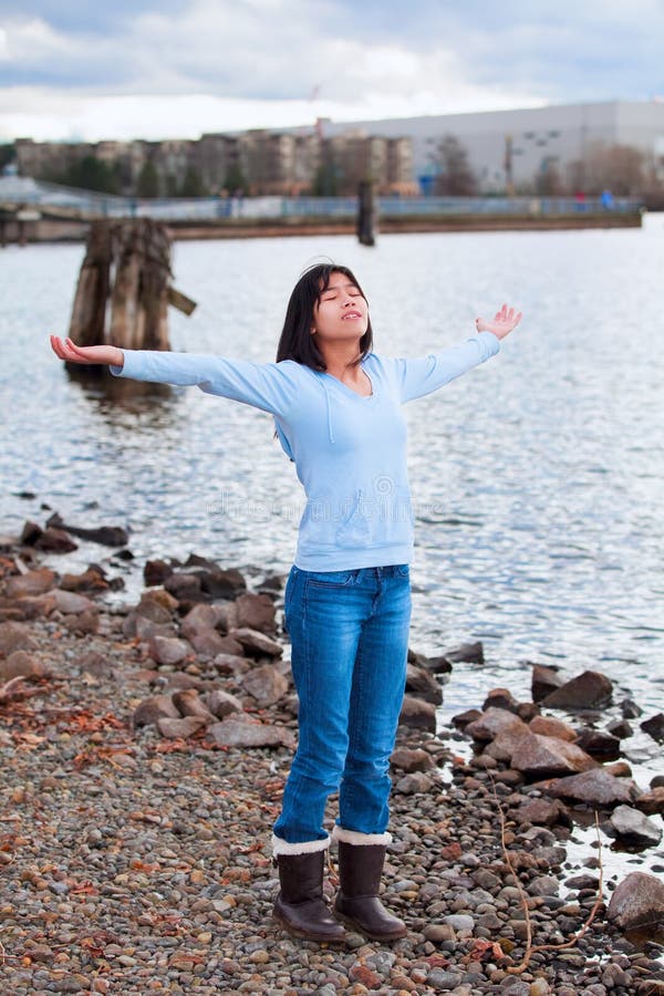 Young biracial teen girl in blue shirt and jeans, arms lifted and outstretched, praising God on rocky shore by lake on cloudy day. Young biracial teen girl in blue shirt and jeans, arms lifted and outstretched, praising God on rocky shore by lake on cloudy day
