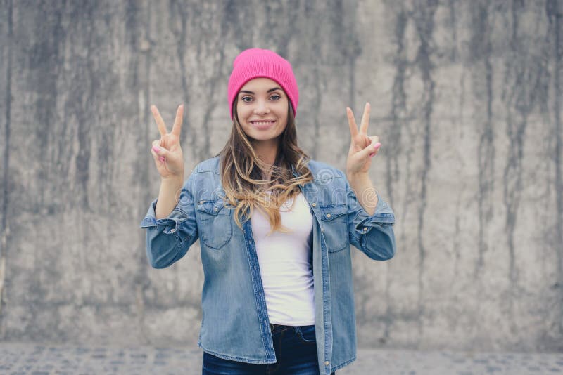 Fille de sourire joyeuse heureuse de hippie dans les vêtements sport et le chapeau rose se tenant contre le mur gris et montrant