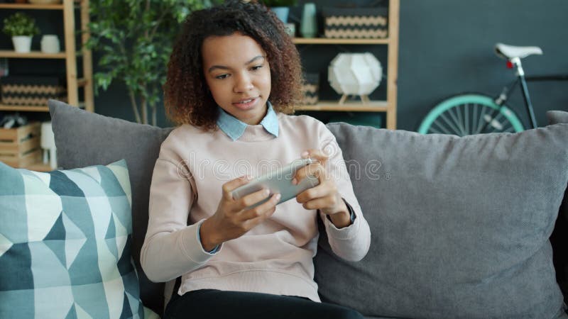 Fille heureuse d'afro-américains jouant l'écran tactile de jeu intelligent de téléphone à la maison en souriant banque de vidéos