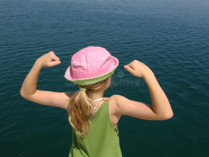 A girl with pink hat and lifting arms, clenched his fist and shows his muscles, biceps, and looking into the clear blue Adriatic sea. Horizontal color photo. A girl with pink hat and lifting arms, clenched his fist and shows his muscles, biceps, and looking into the clear blue Adriatic sea. Horizontal color photo.