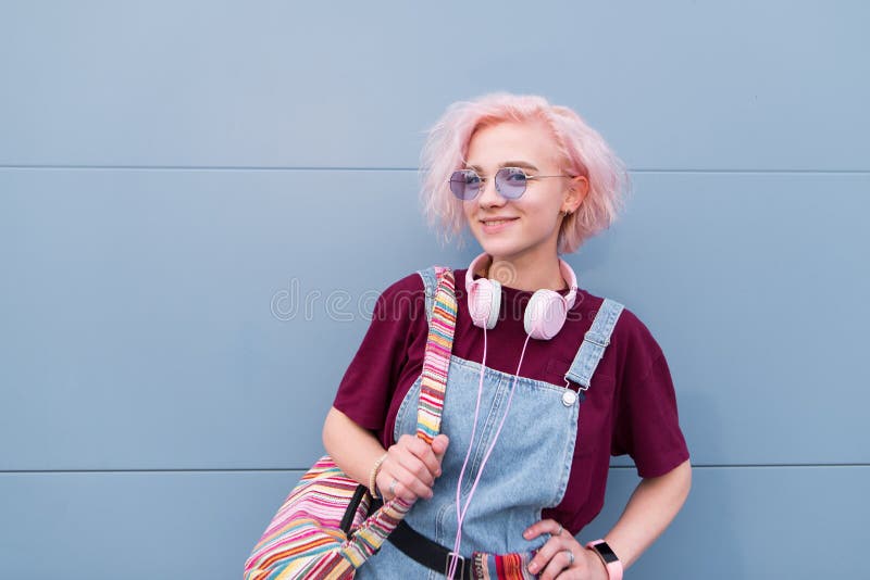 Fille intelligente heureuse avec les cheveux roses se tenant dans la perspective d'un mur bleu examinant la caméra et le sourire photo stock Fille intelligente heureuse avec les cheveux roses se tenant dans la perspective d'un mur bleu examinant la caméra et le sourire photo stock