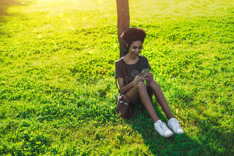 Fille noire gaie avec le téléphone portable se reposant sur l'herbe photo stock Fille noire gaie avec le téléphone portable se reposant sur l'herbe photo stock