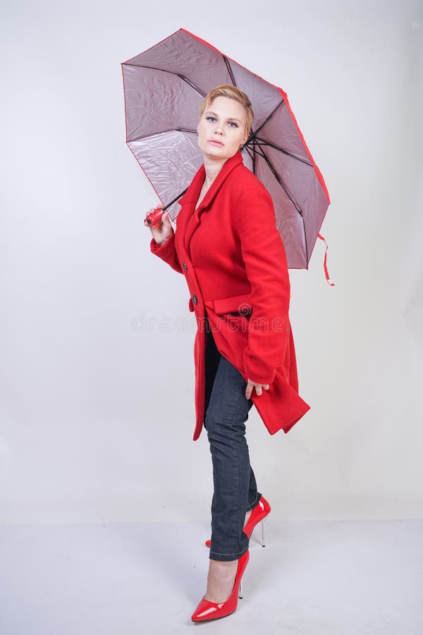Fille potelée mignonne de cheveux courts dans un manteau moderne de laine de ville tenant un parapluie rouge et posant sur un fon images libres de droits Fille potelée mignonne de cheveux courts dans un manteau moderne de laine de ville tenant un parapluie rouge et posant sur un fon images libres de droits