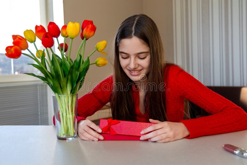 Fille souriante avec cadeau et tulipes. Jeune femme en chandail rouge tenant joyeusement une boîte cadeau rouge près d'un bouquet images libres de droits