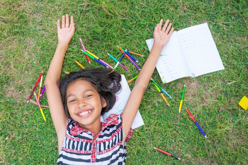 Fille thaïlandaise asiatique se trouvant sur l'herbe avec le crayon et le homew colorés photographie stock libre de droits Fille thaïlandaise asiatique se trouvant sur l'herbe avec le crayon et le homew colorés photographie stock libre de droits