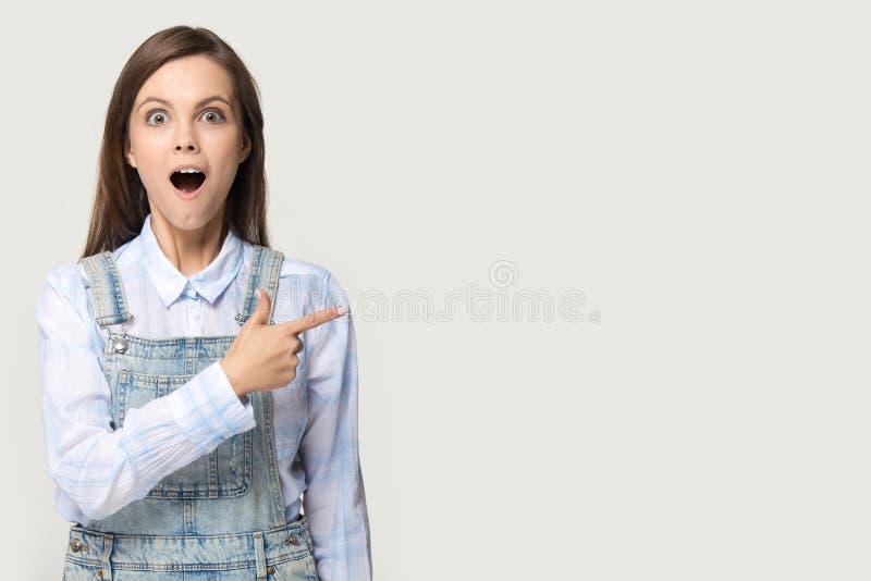 Fille étonnée dirigeant le doigt de côté au fond gris de studio de copyspace photo stock Fille étonnée dirigeant le doigt de côté au fond gris de studio de copyspace photo stock
