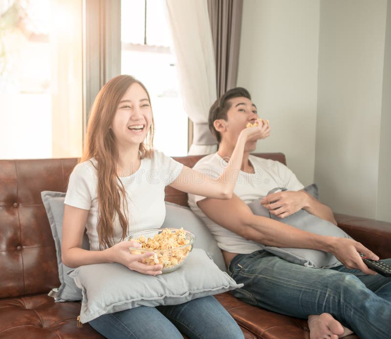 Filme de observação dos pares asiáticos que senta-se em um sofá em casa alegre e na pipoca da tomada ao amigo de menino foto de stock Filme de observação dos pares asiáticos que senta-se em um sofá em casa alegre e na pipoca da tomada ao amigo de menino foto de stock