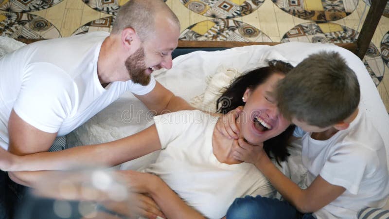 Fils et père chatouillant la mère sur le lit, famille heureuse ayant le temps d'amusement ensemble à la maison