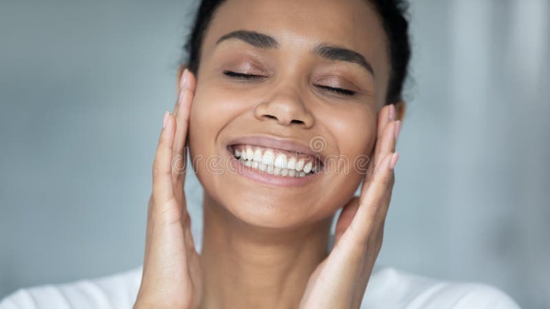 Close up head shot beautiful African American young woman enjoying perfect skin with closed eyes, touching face with hands, attractive girl with healthy smile doing facial massage, skincare procedure. Close up head shot beautiful African American young woman enjoying perfect skin with closed eyes, touching face with hands, attractive girl with healthy smile doing facial massage, skincare procedure