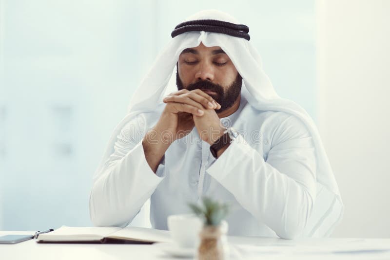 Finding answers through prayer. a young businessman dressed in Islamic traditional clothing praying while working in his stock photography