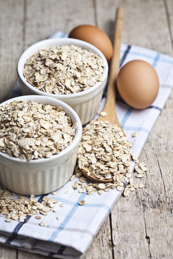 Fiocchi di avena in ciotole ceramiche ed uova fresche di legno e del cucchiaio del pollo sul fondo di legno rustico della tavola fotografia stock Fiocchi di avena in ciotole ceramiche ed uova fresche di legno e del cucchiaio del pollo sul fondo di legno rustico della tavola fotografia stock