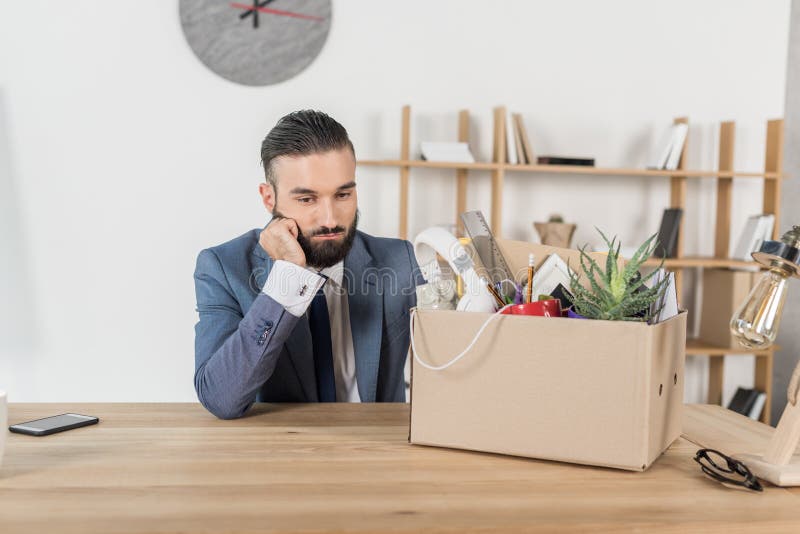 Fired upset businessman sitting at workplace with cardboard box with office supplies. Corporate, unemployment.