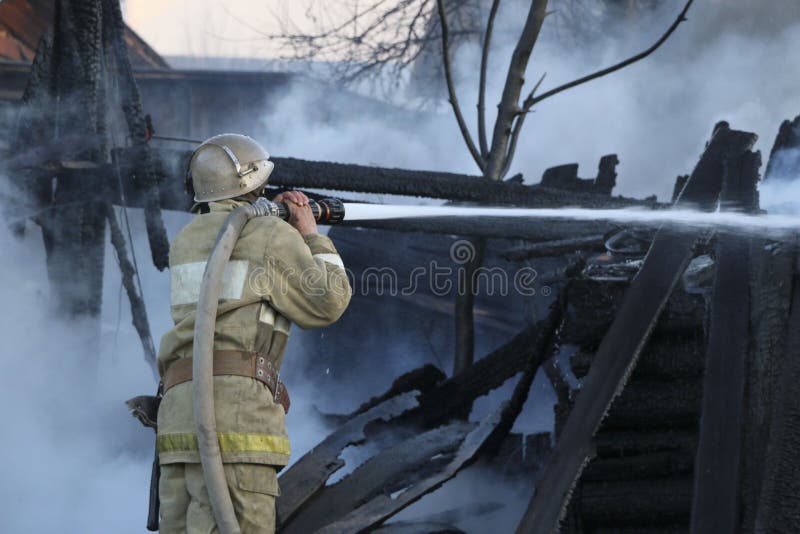 Firefighter extinguishes the fire. Fireman holding a hose with water, watering a strong stream of burning wooden structure in the stock photography Firefighter extinguishes the fire. Fireman holding a hose with water, watering a strong stream of burning wooden structure in the stock photography