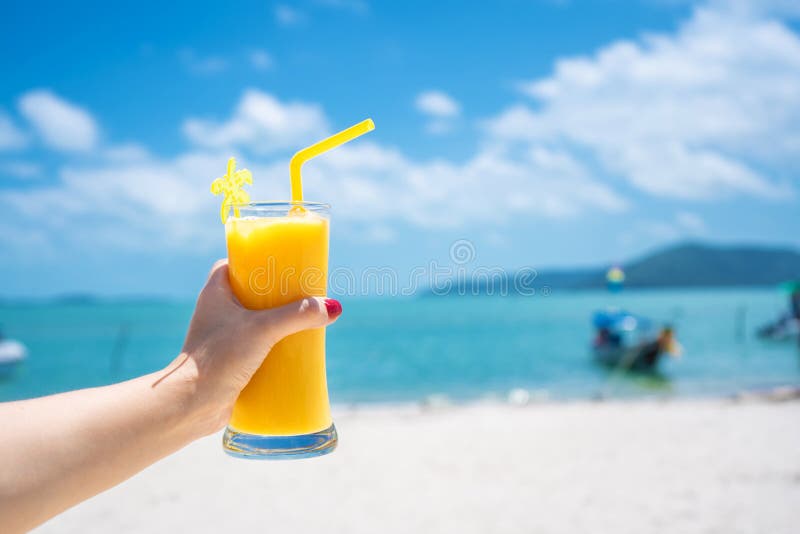 First-person view. Girl holds a glass cup of cold mango fresh on the background of a sandy tropical beach. White sand and a boat.