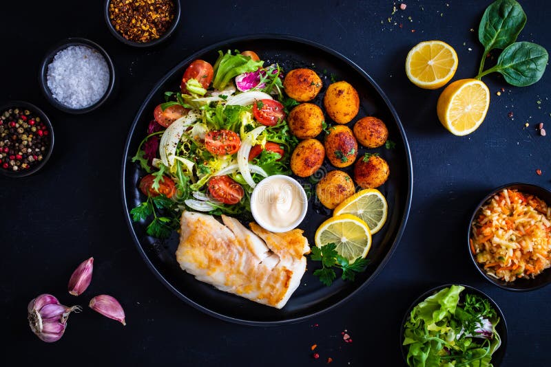 Fish dish - fried cod fillet with potatoes and vegetable salad on black wooden table