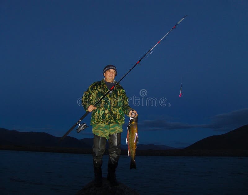 Fisherman standing on lake shore and holding spinning rod and big salmon fish caught at night