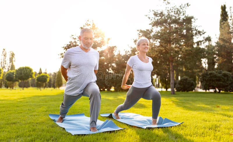 Happy positive fit mature couple of senior woman and man practicing partner yoga on open fresh air outside standing barefoot in warrior Virabhadrasana pose in nature on open air of city park exterior. Happy positive fit mature couple of senior woman and man practicing partner yoga on open fresh air outside standing barefoot in warrior Virabhadrasana pose in nature on open air of city park exterior