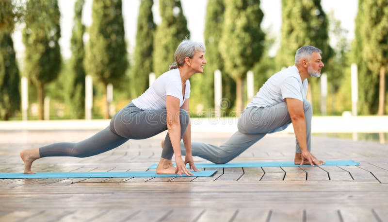 Happy positive fit mature couple of senior woman and man practicing partner yoga on open fresh air outside standing barefoot in warrior Virabhadrasana pose against background of city park exterior. Happy positive fit mature couple of senior woman and man practicing partner yoga on open fresh air outside standing barefoot in warrior Virabhadrasana pose against background of city park exterior