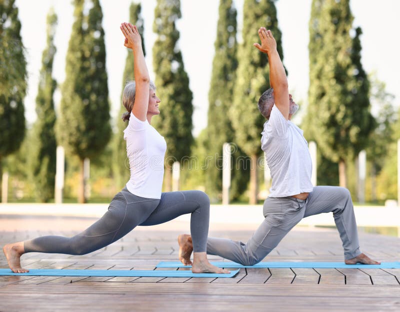 Happy positive fit mature couple of senior woman and man practicing partner yoga on open fresh air outside standing barefoot in warrior Virabhadrasana pose against background of city park exterior. Happy positive fit mature couple of senior woman and man practicing partner yoga on open fresh air outside standing barefoot in warrior Virabhadrasana pose against background of city park exterior