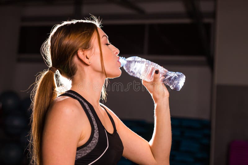 Fitness young woman drinking water in the gym. Muscular woman taking break after exercise