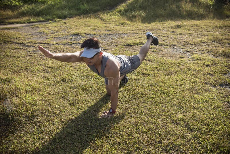 A fit and youthful asian man performs bird dogs on the grass of an open field. Lower back and ab workout. A fit and youthful asian man performs bird dogs on the grass of an open field. Lower back and ab workout.