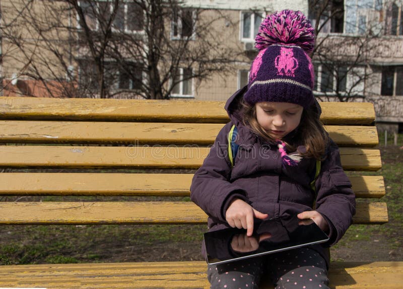 Five years old girl sittion on a bench and touching with finger her digital tablet, outdoors royalty free stock image Five years old girl sittion on a bench and touching with finger her digital tablet, outdoors royalty free stock image