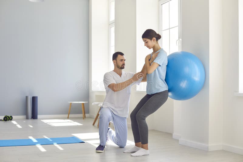 Rehabilitation after injury. Physiotherapist helping women with backache to restore spinal health after physical trauma. Young female athlete doing back exercise using soft yoga ball. Rehabilitation after injury. Physiotherapist helping women with backache to restore spinal health after physical trauma. Young female athlete doing back exercise using soft yoga ball