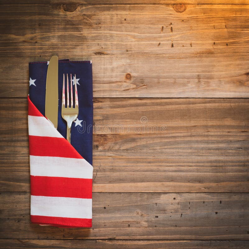 Fourth of July Table Place Setting with a fork, knife and flag napkin in warm light on rustic wood board background with room or space for copy, text, or your words.  It`s a square photo with above view. Fourth of July Table Place Setting with a fork, knife and flag napkin in warm light on rustic wood board background with room or space for copy, text, or your words.  It`s a square photo with above view