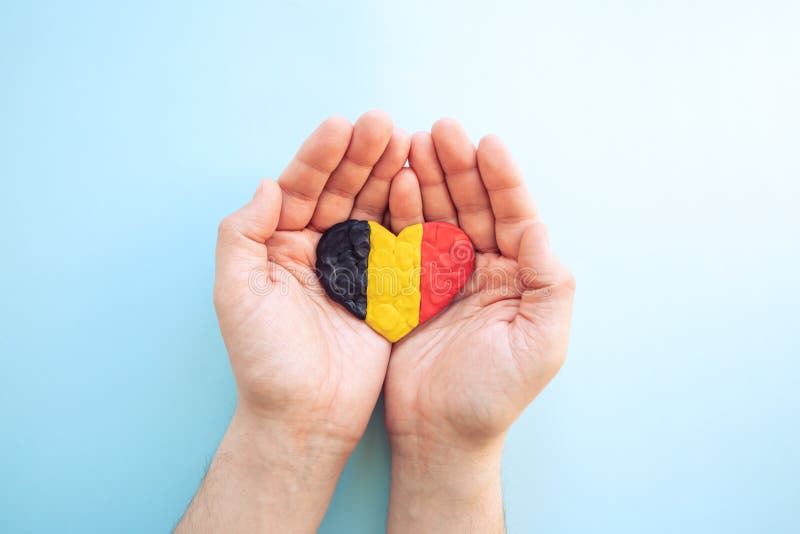 Flag of Belgium. Heart shape of black, yellow and red plasticine modeling clay in male hands on blue background. Belgian flag
