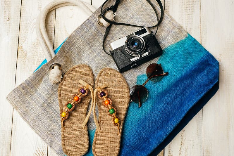 Flat lay of summer fashion with camera, slippers, sunglasses and other girl accessories on top of the bag on white wooden backgrou