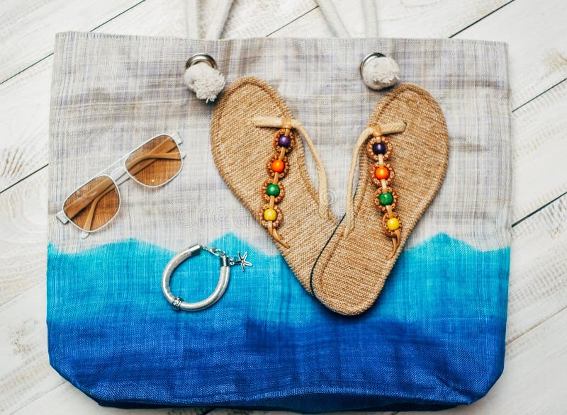 Flat lay of summer fashion with slippers and sunglasses on top of the bag on white wooden background