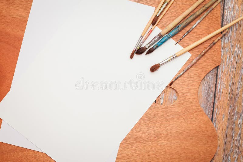 Flat lay of white Papers, paint brushes and wooden palette on brown wooden table.