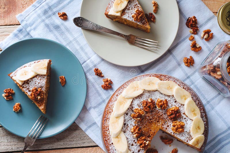 Flatlay with sliced banana cake with powdered sugar and walnut on plate with fork and glass jar full of nuts. Cozy top view in light tones on wooden background with checked kitchen towel. Vertical flatlay.