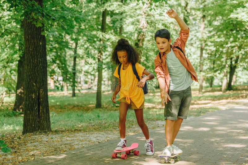 Flerkulturella skolbarn rider skateboardbord i grön solopark royaltyfri foto