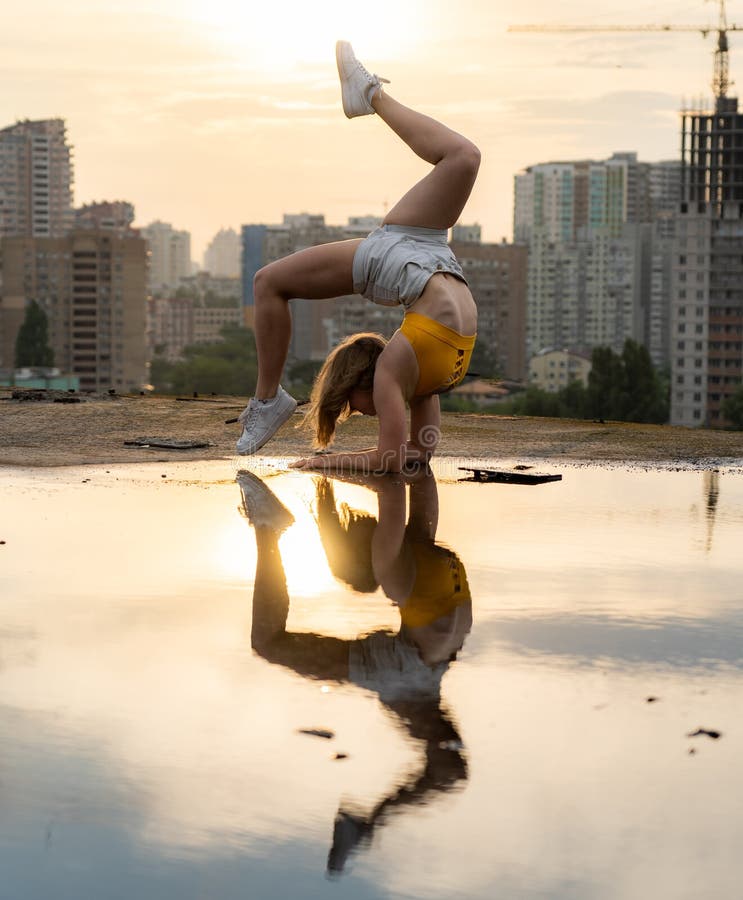 Flexible female gymnast doing handstand and calisthenic with reflection in the water on cityscape background during