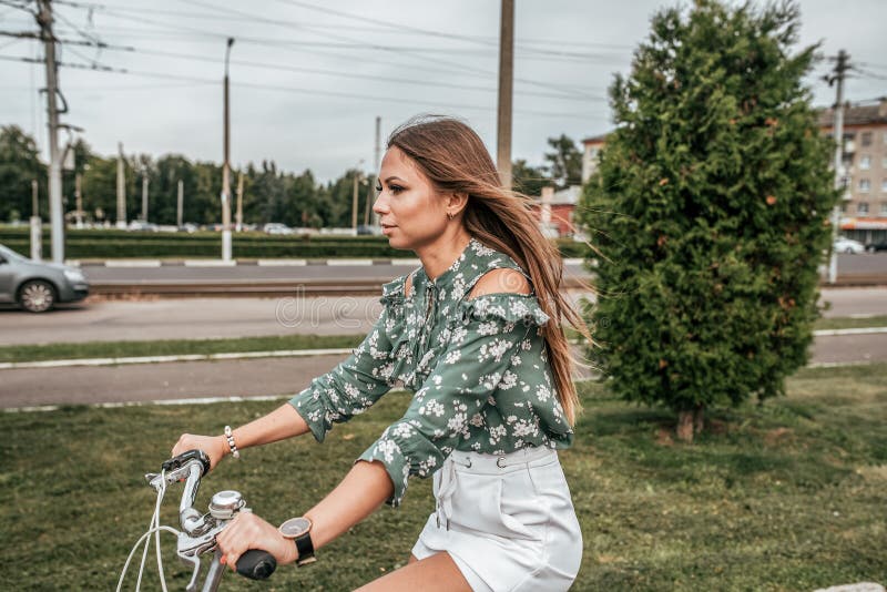 Flicka i sommaren i staden som rider en cykel, mot bakgrunden av träd och gröna bilar I en grön tröja och vit arkivfoto Flicka i sommaren i staden som rider en cykel, mot bakgrunden av träd och gröna bilar I en grön tröja och vit arkivfoto