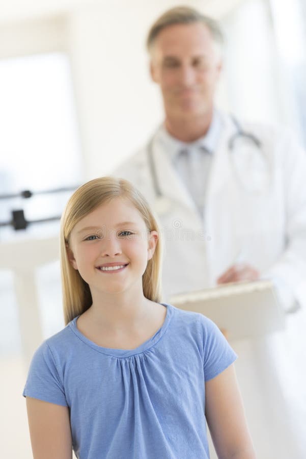 Flicka som ler med doktor Standing In Background på kliniken royaltyfria foton Flicka som ler med doktor Standing In Background på kliniken royaltyfria foton