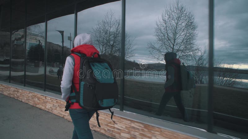 Flickan i den röda, sömnlösa jackan går bredvid de stora fönstren på grumlig dag