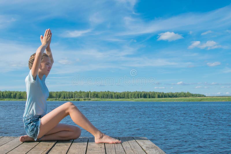 Flickan som sitter på pir, i en yoga, poserar, på bakgrunden av naturen arkivbild Flickan som sitter på pir, i en yoga, poserar, på bakgrunden av naturen arkivbild