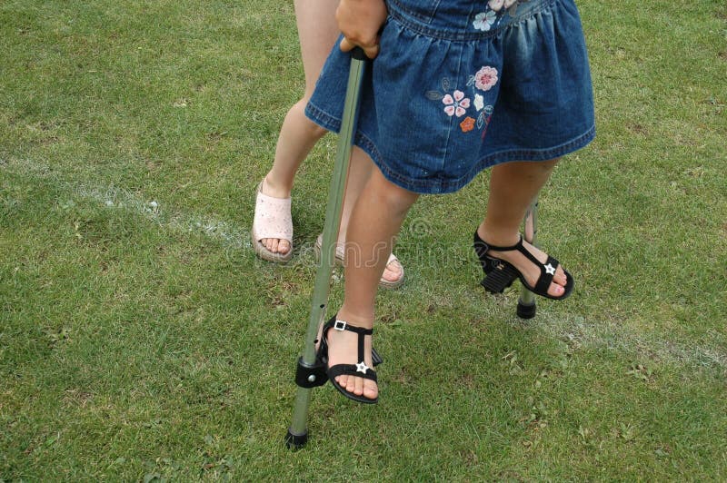 One girl balances on a pair of stilts, will the second holds her to get the balance correct. On grass with a faint white line across the shot. One girl balances on a pair of stilts, will the second holds her to get the balance correct. On grass with a faint white line across the shot.