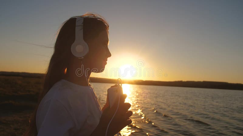 Flickatonåring i hörlurar med near vatten för telefon på solnedgången långsam rörelse