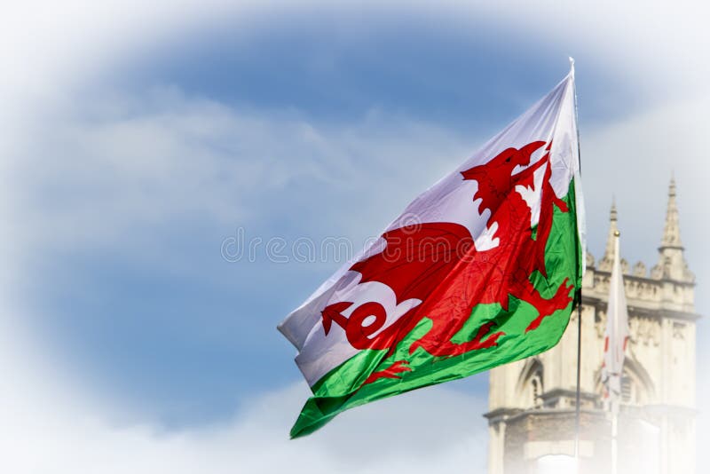 Welsh Dragon Flag flying in blue cloudy sky with the top of a castle like building in the background. Welsh Dragon Flag flying in blue cloudy sky with the top of a castle like building in the background.
