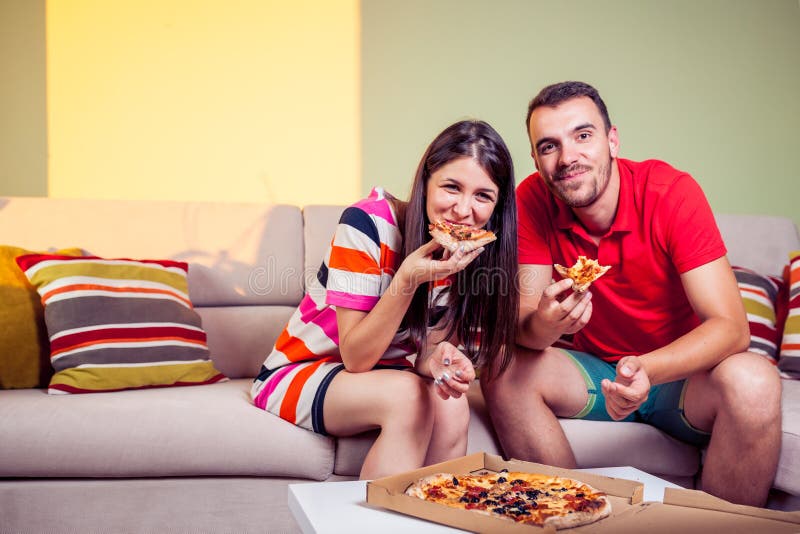 Flippige junge Paare, die Pizza auf einer Couch essen