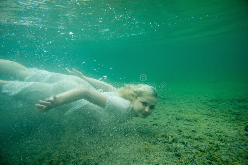 A floating woman. Underwater portrait. Girl in white dress swimming in the lake. Green marine plants, water