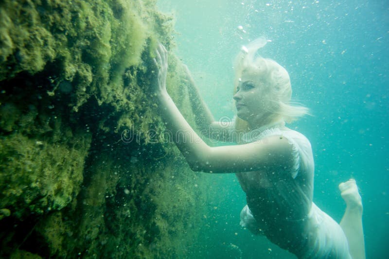 A floating woman. Underwater portrait. Girl in white dress swimming in the lake. Green marine plants, water