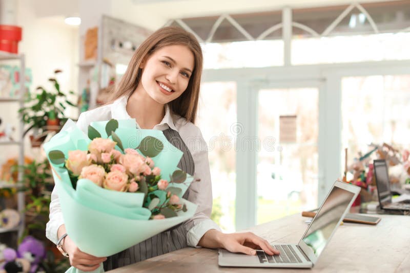 Florista de sexo femenino joven con el ramo y ordenador portátil que trabaja en floristería fotos de archivo libres de regalías Florista de sexo femenino joven con el ramo y ordenador portátil que trabaja en floristería fotos de archivo libres de regalías