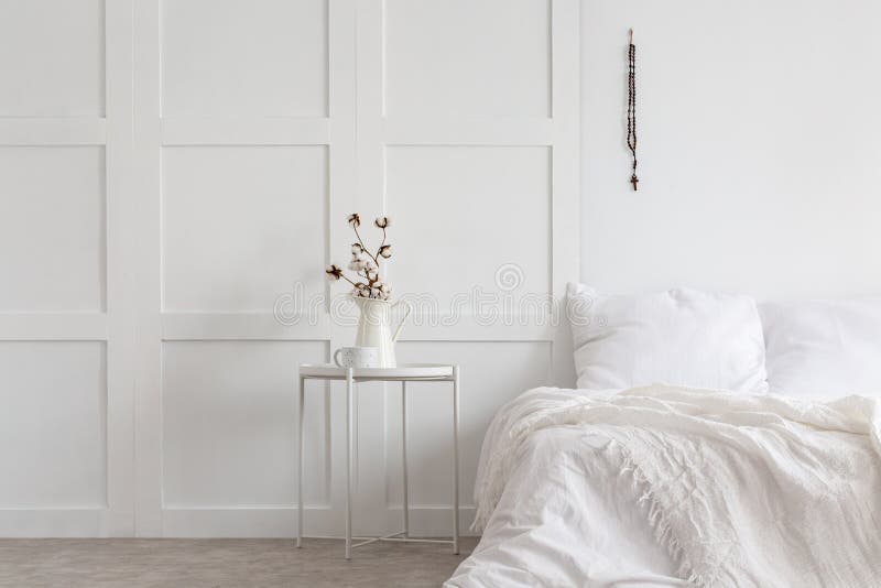 Flowers on table next to bed with pillows and sheets in white minimal bedroom interior