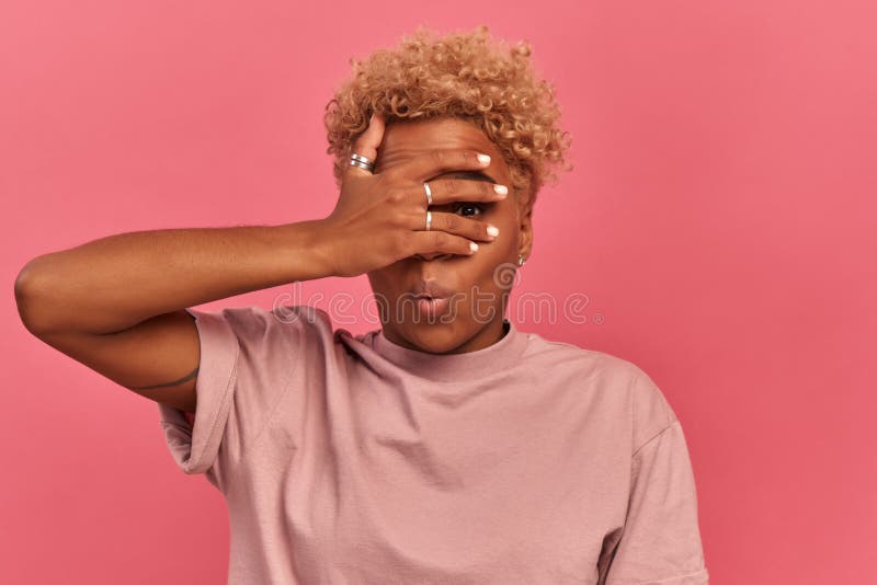 Omg, I dont want to see it. Young African American woman peeks through fingers covering face with hand embarrassed female peeping while standing against pink background with place for text. Omg, I dont want to see it. Young African American woman peeks through fingers covering face with hand embarrassed female peeping while standing against pink background with place for text.