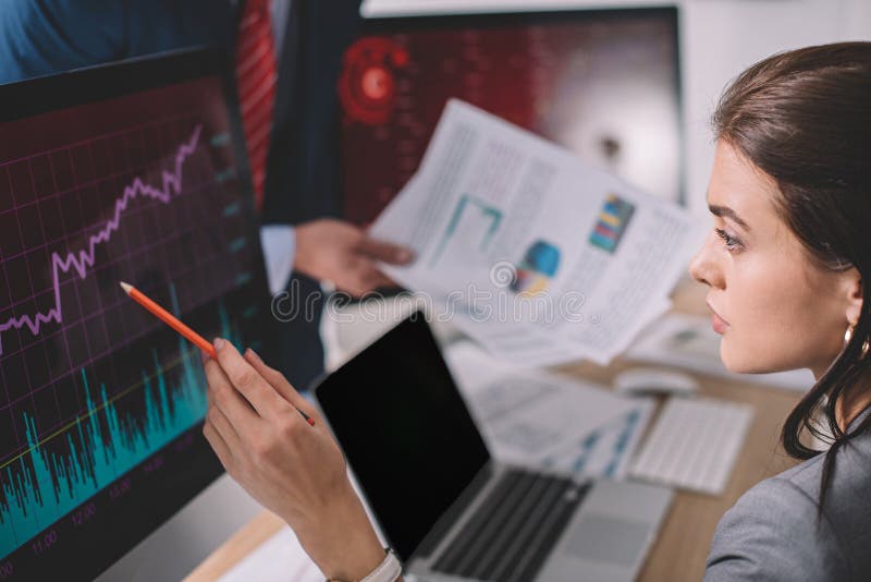 Focus of information security analyst pointing on charts on computer monitor near colleague with papers on grey stock images