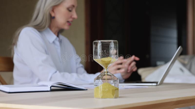 Focus op uurglas op achtergrond oudere zakenvrouw die werkt op computer